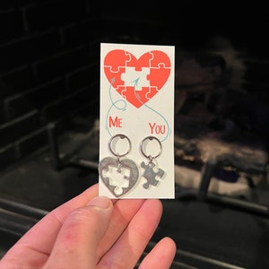 May include: A pair of silver earrings with puzzle piece charms. The charms are shaped like a heart with a missing piece. The card behind the earrings says "Me" and "You" with arrows pointing to the missing piece of the puzzle.