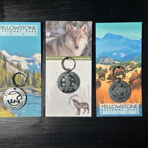 May include: Three keychains featuring Yellowstone National Park. The keychains are silver with a black and white image of a bear, a wolf, and a bison. The keychains are displayed on cards with images of the animals and the text "Yellowstone National Park" and the animal name.