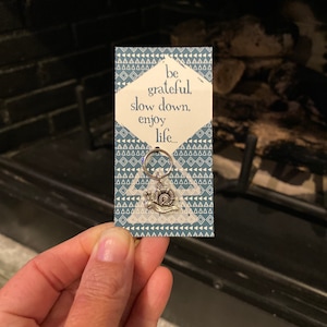 May include: A small card with a blue and white geometric pattern. The card has a silver snail charm attached to it. The card says "be grateful, slow down, enjoy life..."