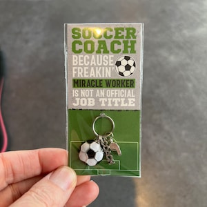 May include: A silver keychain with a soccer ball charm, a whistle charm, and a charm that says "coach". The keychain is attached to a card that says "Soccer Coach because freakin' miracle worker is not an official job title".