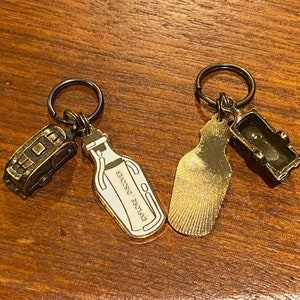 May include: Two gold-toned keychains with charms. One keychain has a charm shaped like a bottle with the text "Explore Discover" on it. The other keychain has a charm shaped like a small bus and a charm shaped like a bottle.