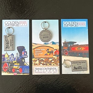 May include: Three keychains featuring images of trains and the Golden Spike National Historical Park. The keychains are displayed on cards with the park's logo and the text "Golden Spike National Historical Park" and "Transcontinental Railroad Promontory, Utah 1869".