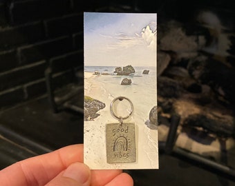 May include: Silver keychain with a rectangular tag that says "good vibes" with a rainbow graphic. The keychain is held in a hand against a blurry background of a beach scene.