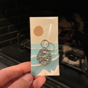 May include: A silver charm with the text "may you always have a shell in your pocket & sand between your toes" on a white card with a blue and white pattern.