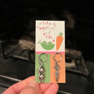 May include: A card with the text "We belong together just like..." and two peas in a pod and a carrot with a smiley face.  The card is attached to two silver charms, one of a pea pod and one of a carrot.