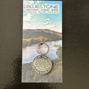 May include: A silver keychain with a round metal charm featuring the words "Bluestone National Scenic River" and a depiction of a canoe on a river. The keychain is displayed on a card with a scenic image of a river and the words "Bluestone National Scenic River Great Blue Heron."
