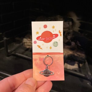 May include: A silver charm with a planet Saturn design, attached to a silver key ring. The charm is on a white card with a pink and orange watercolor background. The card has the text "You're Stellar" in black lettering.