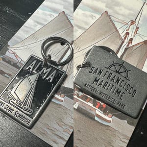 May include: Two silver keychains with a black background. The first keychain features a sailboat with the text "ALMA 91 SCOW SCHOONER". The second keychain features the text "SAN FRANCISCO MARITIME NATIONAL HISTORICAL PARK".