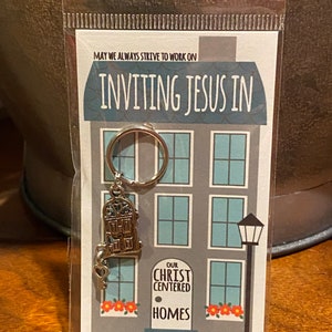 May include: A silver key chain with a house charm and a key. The charm has a door with the words "Our Christ Centered Homes" on it. The card says "May we always strive to work on inviting Jesus in. Home sweet home!"