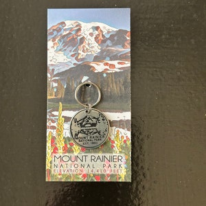 May include: A silver keychain with a round metal charm featuring a black and white image of Mount Rainier National Park. The charm has the text "Mount Rainier National Park Est. 1899". The keychain is attached to a card with a colorful image of Mount Rainier and the text "Mount Rainier National Park Elevation 14,410 feet."
