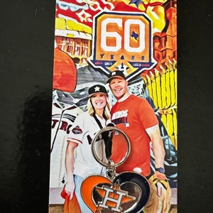 May include: A couple poses in front of a mural celebrating 60 years of the Houston Astros. The woman is wearing a white and blue Astros jersey and the man is wearing an orange and blue Astros jersey. They are holding a silver keychain with a heart-shaped charm featuring the Astros logo.