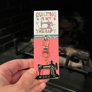 May include: A black and gold sewing machine keychain with the text "#ISEW" on the bottom. The keychain is attached to a pink and white card with the text "QUILTING IS MY THERAPY".