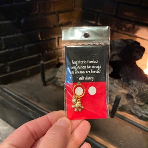 May include: A red card with a white quote by Walt Disney: "laughter is timeless imagination has no age and dreams are forever."  A gold charm with a heart and a Mickey Mouse hand is attached to the card.