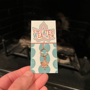 May include: A gold-colored maple leaf charm on a white card with blue polka dots. The card has the text "SWEATER WEATHER" in a stylized font.
