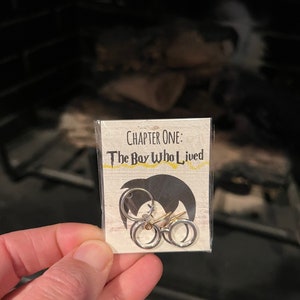 May include: A close-up of a small card with the text "CHAPTER ONE: The Boy Who Lived" printed on it. Two silver hoop earrings with a small lightning bolt charm are displayed on the card.