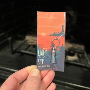 May include: A silver keychain with a lighthouse charm and a card that says "be a light for all to see, matthew 5:16". The card has a sunset scene with a lighthouse on a cliff.