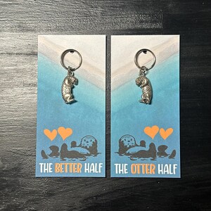 May include: Two silver keychains with a small otter charm. The keychains are on cards with a blue and white background. The cards have the text "The Better Half" and "The Otter Half" with two orange hearts above each otter.