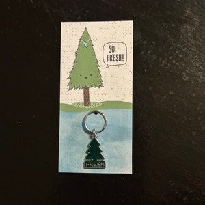 May include: A green pine tree keychain with the word "FRESH" on it. The keychain is attached to a card with a cartoon pine tree and the words "SO FRESH!" in a speech bubble.