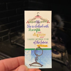 May include: A white card with a floral design and a silver hanger charm. The card has the text "She is clothed with strength and dignity and laughs without fear of the future. Proverbs 31."