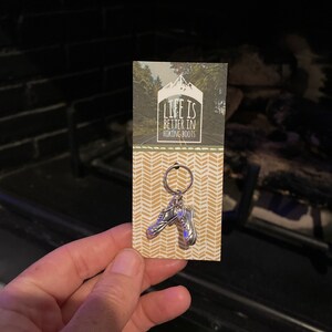 May include: A silver hiking boot keyring with a key ring. The keyring is attached to a card with a chevron pattern and the text "Life is better in hiking boots".