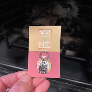 May include: A pink and yellow card with the text "ME VS. ME" and a silver alarm clock charm attached to a silver ring.