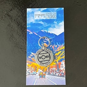 May include: Silver keychain with a round tag featuring the state of Colorado logo and the text "Colorado Est. 1876". The keychain is displayed on a card with a mountain scene and the text "Established State of 1876 Colorado".