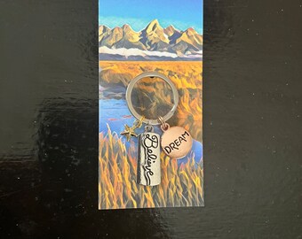 May include: A silver keychain with a charm that says "Believe" and a round charm that says "Dream". The charms are attached to a silver key ring. The keychain is on a card with a picture of mountains and a field.