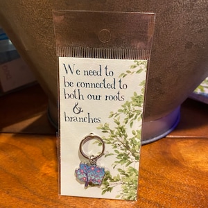 May include: A white card with the text "We need to be connected to both our roots & branches."  A silver key chain with a blue and pink tree charm is attached to the card.
