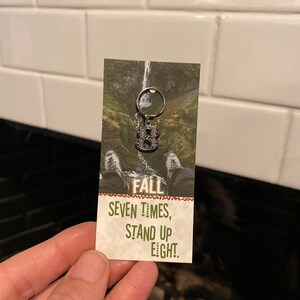 May include: A silver keychain with the number eight on it. The keychain is attached to a card with the text "Fall seven times, stand up eight."