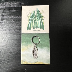 May include: A silver keychain with a tree design. The keychain is on a card with a watercolor painting of a forest and the words "Simplify" and "Life is Good".