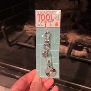May include: A silver keyring with a turquoise and silver pizza cutter charm. The keyring is packaged on a blue and white card with the text "Sharpest Tool in the Kit".