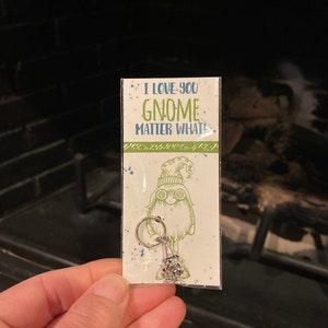 May include: A white card with green and blue text that reads "I love you gnome matter what." A silver gnome charm is attached to the card with a silver ring.