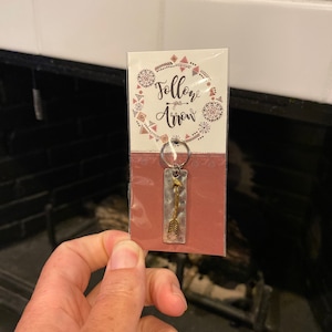 Könnte beinhalten: Ein silberner und goldener Schlüsselanhänger mit einem kleinen Pfeil-Anhänger. Der Schlüsselanhänger ist auf einer rosa Karte verpackt mit dem Text "Follow your arrow" in einem weißen Kreis mit einem geometrischen Muster.