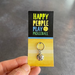 May include: A silver pickleball charm keyring with a black and yellow card that reads "Happy People Play Pickleball".