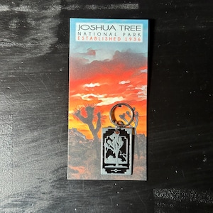 May include: A rectangular keychain featuring a Joshua Tree National Park design. The keychain has a silver-colored metal frame with a black and white image of a Joshua tree. The background card displays the park's name and establishment year against a sunset backdrop.