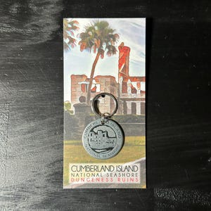 May include: A silver keychain with a round charm featuring a black and white illustration of a building with the text "Cumberland Island National Seashore". The keychain is attached to a card with the text "Cumberland Island National Seashore Dungeness Ruins".