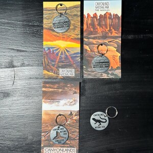May include: Four different keychains featuring the Canyonlands National Park logo. Each keychain has a different design, including a lizard, a sunset, and a rock formation. The keychains are made of metal and have a silver finish.