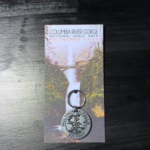 May include: A keychain featuring a silver-colored Multnomah Falls medallion with the text "MULTNOMAH FALLS 621 FEET" and a scenic card with the text "COLUMBIA RIVER GORGE NATIONAL SCENIC AREA MULTNOMAH FALLS".