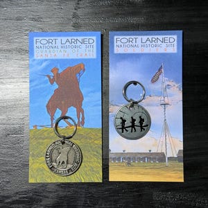 May include: Two keyrings featuring the Fort Larned National Historic Site. One keyring has a circular pendant with a depiction of soldiers, the other has a pendant with a rider on a horse. Both keyrings are attached to cards with the site's name.