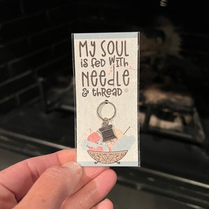 May include: A silver charm with a spool of thread and needles, attached to a key ring. The charm is displayed on a card with the text "My soul is fed with needle & thread".