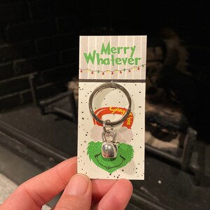 May include: A silver keychain with a bell charm and a green Grinch face. The keychain is attached to a card that says "Merry Whatever" with a string of lights above the text.