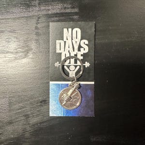 May include: Silver keychain with a weight plate charm and a barbell with a figure lifting it. The keychain has a black card attached with the text "NO DAYS OFF".