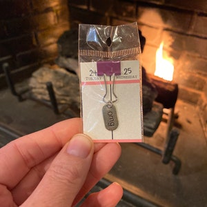 May include: A silver charm with the word "omg" on it. The charm is attached to a silver ring and is packaged in a clear plastic bag with a white card that says "24 Tuesday" and "25 Wednesday".