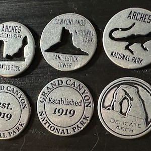 May include: Six silver-colored metal tokens featuring designs and text related to U.S. National Parks. The tokens include images of rock formations and the names of parks like Arches and Grand Canyon. Some tokens have the text "Established 1919".