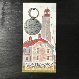 Könnte beinhalten: Ein silberner Schlüsselanhänger mit einem runden Metallanhänger, der einen Vogel und den Text "Gateway National Recreation Area" zeigt. Der Anhänger ist an einem silbernen Schlüsselring befestigt. Das Hintergrundbild zeigt einen Leuchtturm und ein Haus.
