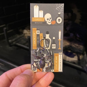 May include: A silver spider charm with a black background card featuring a skull, candles, and books with titles like "Curses & Disguises", "Alchemy", "Poisons", "Black Magic", "Spells & Incantations", and "Book of Spells".