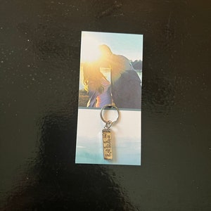 May include: A silver keychain with a rectangular charm. The charm has the words "It is well with my soul" in a handwritten font. The keychain is attached to a card with a photo of a couple at sunset.