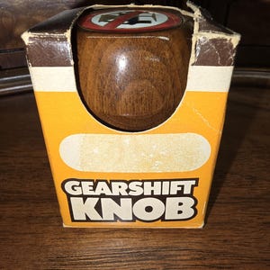 Puede incluir: Una perilla de cambio de marchas de madera marrón con una señal de prohibido estacionar blanca y roja en la parte superior. La perilla está en una caja amarilla y marrón con las palabras "Gearshift Knob" impresas en la parte delantera.