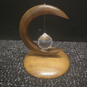 Peut inclure: Une sculpture en forme de croissant de lune en bois, avec une base ronde, tenant une sphère de cristal facettée. Le bois est de couleur marron chaud et le cristal est transparent. La sculpture est conçue pour suspendre le cristal au centre.