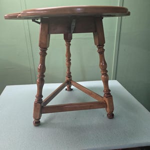 May include: A small, antique wooden table with a round top and three ornate legs. The table is made of light brown wood and features a triangular support structure. The table's top is approximately 24 inches in diameter.